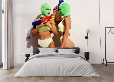 Two beautiful sexy women in green underwear. Models wearing bandit balaclava mask. Hot seductive female in nice lingerie posing in the street at sunny day, blue sky. Crime and violence. Hold water gun Wall mural