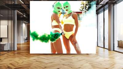 Two beautiful sexy women in green underwear. Models wearing bandit balaclava mask. Hot seductive female in nice lingerie posing in the street at sunny day. Crime and violence. Hold green smoke bomb Wall mural