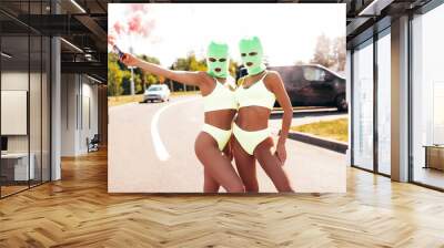 Two beautiful sexy women in green underwear. Models wearing bandit balaclava mask. Hot seductive female in nice lingerie posing in the street at sunny day. Crime and violence. Hold smoke bomb Wall mural