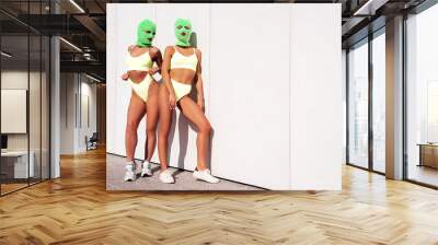 Two beautiful sexy women in green underwear. Models wearing bandit balaclava mask. Hot seductive female in nice lingerie posing in the street at sunny day. Crime and violence. Perfect body. Near wall Wall mural