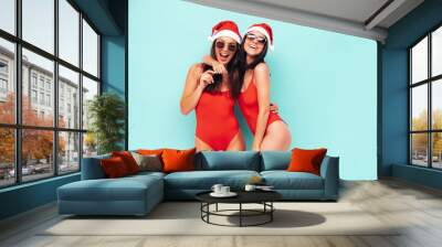 Two beautiful women celebrating New Year. Happy gorgeous female in red sexy body. Having fun at New Year's Eve party. Holiday celebration. Charming models in Santa Claus hats laughing near blue wall Wall mural