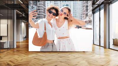 Two young beautiful smiling cute women in trendy summer sundress. Sexy carefree models posing in the street in sunny day. Positive models having fun and hugging. Cheerful and happy, take selfie Wall mural