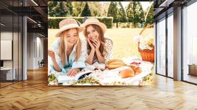 Two young beautiful smiling female in trendy summer sundress and hats.Carefree women making picnic outside.Positive models sitting on plaid on grass, reading book, eating fruits and cheese Wall mural