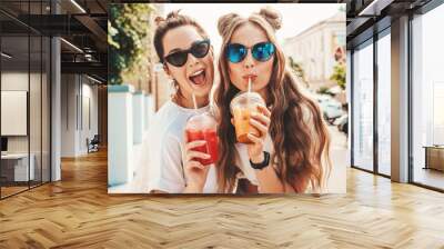 Two young beautiful smiling hipster female in trendy summer clothes. Carefree women posing outdoors.Positive models holding and drinking fresh cocktail smoothie drink in plastic cup with straw Wall mural