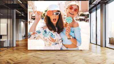 Two young beautiful smiling hipster girls in trendy summer clothes and panama hat. Sexy carefree women posing on street background.Positive models having fun in sunglasses.Eating candy lollipop Wall mural