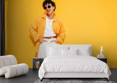 Young attractive man with short curly hair hairstyle. Smiling handsome male in casual stylish shirt and shorts clothes posing in studio, isolated on yellow. Cheerful and happy model, sunglasses Wall mural