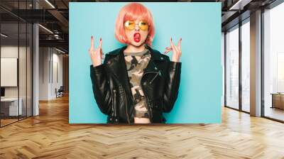 Young beautiful hipster bad girl in black leather jacket and earring in nose.Sexy carefree  woman posing in studio in pink wig near blue wall.Confident model in sunglasses.Shows rock and roll sign Wall mural