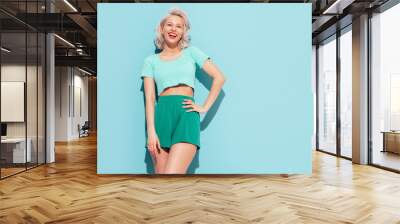 Young beautiful smiling female in trendy summer shorts and top clothes. Sexy carefree woman posing near blue wall in studio. Positive blond model having fun and going crazy. Cheerful and happy Wall mural