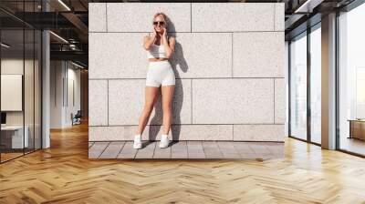 Young beautiful smiling female in trendy summer white cycling shorts and tank top clothes. Sexy carefree woman posing in street. Positive model . Cheerful and happy.  In sunglasses, near wall Wall mural