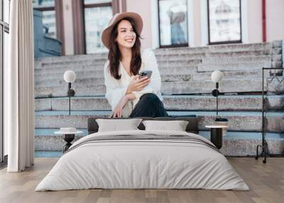 Young beautiful smiling woman in trendy summer clothes. Carefree female posing in street in sunny day. Positive model holds smartphone, looks at cellphone screen, uses mobile apps, in hat Wall mural