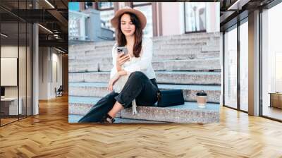 Young beautiful smiling woman in trendy summer clothes. Carefree female posing in street in sunny day. Positive model holds smartphone, looks at cellphone screen, uses mobile apps, in hat Wall mural