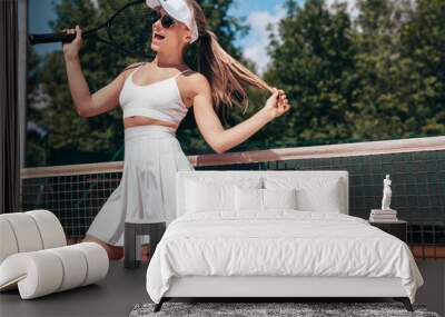 Young beautiful smiling woman wearing fashionable white dress skirt and tank top. Hot model posing on tennis court at summer sunny day. Sexy female stands near net, cheerful and happy, sunglasses Wall mural