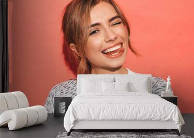 Young beautiful woman looking at camera. Trendy girl in casual summer shirt clothes with natural makeup. Positive female smiling. Funny model posing near pink wall in studio.Shows tongue and winks Wall mural