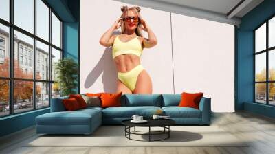 Young smiling blond model in summer swimwear yellow bathing suit. Sexy carefree woman having fun and going crazy. Female posing in the street in sunny day. Cheerful and happy. Near wall, sunglasses Wall mural