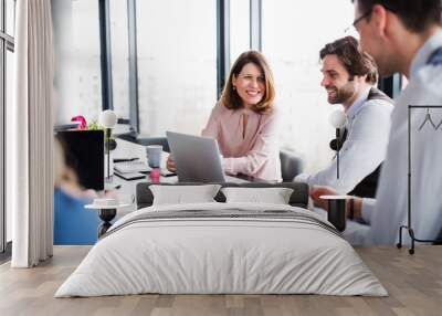 A group of young business people with laptop sitting in an office, talking. Wall mural