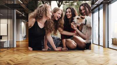 A group of young friends with a dog sitting on grass on a roadtrip through countryside. Wall mural