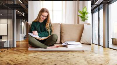 A happy young female student sitting on sofa, studying. Wall mural