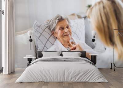 A health visitor examining a sick senior woman lying in bed at home with stethoscope. Wall mural