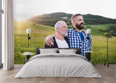 A portrait of an adult hipster son with senior father in nature at sunset, arms around each other. Wall mural