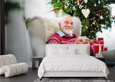 A portrait of senior man with a cup sitting on armchair at home at Christmas time. Wall mural