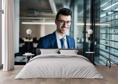 A portrait of young businessman standing in an office, looking at camera. Wall mural