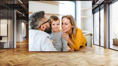 A portrait of young family with a toddler girl indoors in kitchen, kissing. Wall mural