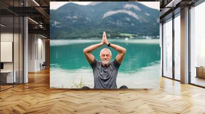 A senior man pensioner sitting by lake in nature, doing yoga exercise. Wall mural