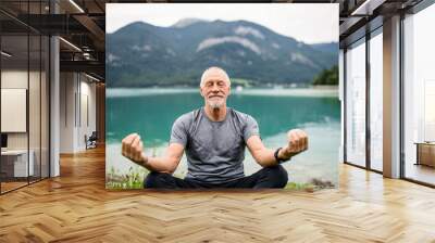 A senior man pensioner sitting by lake in nature, doing yoga exercise. Wall mural