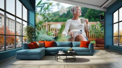 A senior woman with a dog and coffee sitting outdoors on a terrace in summer. Wall mural