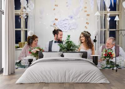 A young couple with parents sitting at a table on a wedding, looking at each other. Wall mural