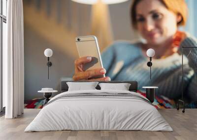 A young woman sitting indoors on a sofa at home, using smartphone. Wall mural