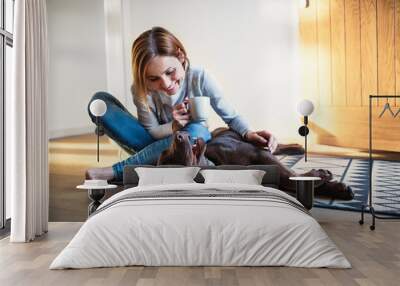 A young woman sitting indoors on the floor at home, playing with a dog. Wall mural