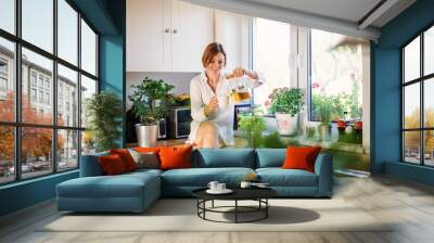 A young woman sitting on worktop indoors in kitchen, pouring tea. Wall mural