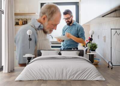 Adult hipster son and senior father indoors in kitchen at home, using tablet. Wall mural