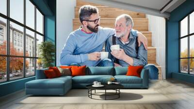 An adult hipster son and senior father sitting on stairs indoors at home, talking. Wall mural