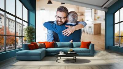 An adult son and senior father indoors at home, hugging. Wall mural