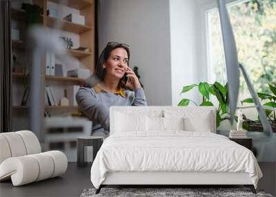 Attractive businesswoman sitting at the desk indoors in office, using smartphone. Wall mural