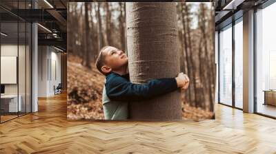 Cute boy on walk in forest, hugging tree, enjoying the peaceful moment, looking up at tree crown. Child connected with nature. Wall mural