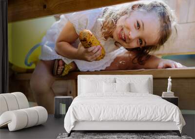 Cute little girl eating grilled corn and looking under the table at camera during garden party. Wall mural