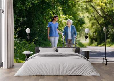 Female caregiver and senior woman with walker on walk in nature. Nurse and elderly woman enjoying a warm day in nursing home, public park. Wall mural