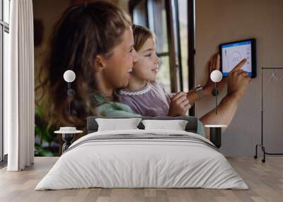 Girl helping mother to adjust, lower heating temperature on thermostat. Concept of sustainable, efficient, and smart technology in home heating and thermostats. Wall mural