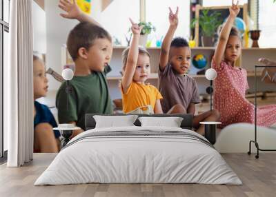 Group of small nursery school children sitting on floor indoors in classroom, raising hands. Wall mural