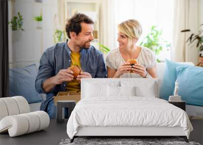 Happy couple sitting on sofa indoors at home, eating hamburgers. Wall mural