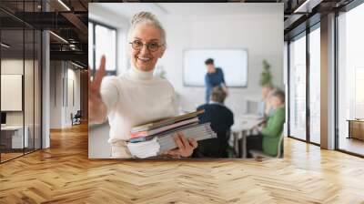 Happy senior woman student with books raising hand and looking at camera in classroom. Wall mural