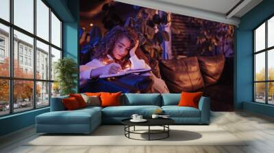 Happy young woman sitting on sofa and writing in diary in the evening in cozy hyyge living room. Wall mural