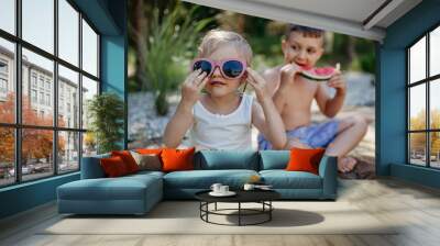 Little chidren sitting near lake and eating watermelon on hot sunny day during summer vacation. Wall mural