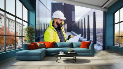 Man engineer standing on construction site, holding blueprints. Wall mural