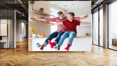 Mature father with small son sitting on kitchen counter indoors, having fun. Wall mural