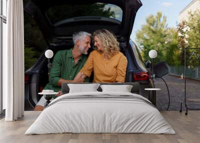 Middle-aged couple sitting in trunk while waiting for charging car before travelling on summer holiday. Wall mural