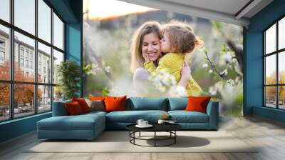 Mother with small daughter standing outdoors in orchard in spring, kissing. Wall mural